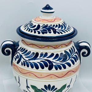 May include: A white ceramic jar with a lid, adorned with blue floral patterns and accents. The jar features two handles and a decorative design with pink and yellow details. The lid is topped with a small, blue finial.