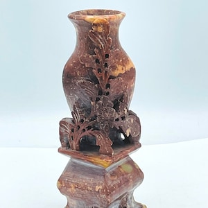 May include: A carved stone vase with a floral design. The vase is brown, red, and white with a square base and four legs.
