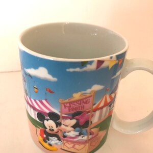 Puede incluir: Una taza de cer&aacute;mica blanca con una imagen de dibujos animados de Mickey Mouse y Minnie Mouse bes&aacute;ndose en un puesto de besos de carnaval. La taza tiene un fondo de cielo azul con nubes blancas y carpas de carnaval de colores.