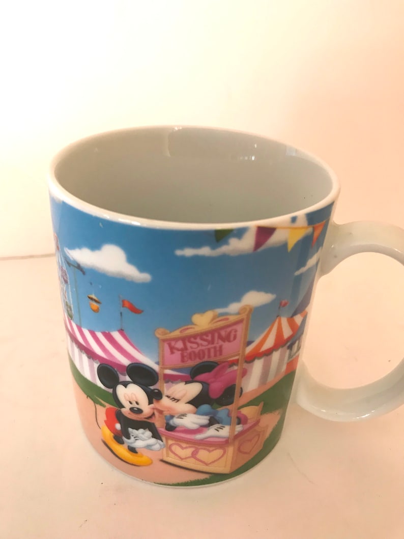 Puede incluir: Una taza de cer&aacute;mica blanca con una imagen de dibujos animados de Mickey Mouse y Minnie Mouse bes&aacute;ndose en un puesto de besos de carnaval. La taza tiene un fondo de cielo azul con nubes blancas y carpas de carnaval de colores.