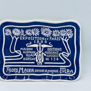 May include: A rectangular ceramic tray with a wavy edge, featuring a deep blue background and white raised relief details. The design includes floral motifs at the top and text reading EXPOSITION DE PARIS 1900, MAISON ALLEMANDE, and ALOYS MAIER.