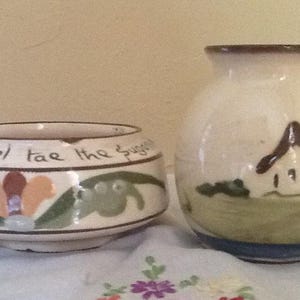 May include: Two ceramic vases. The vase on the left is a small, round bowl with a floral design and the text "versel tae the suga". The vase on the right is a small, round vase with a house and tree design.