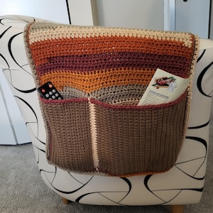 May include: A handmade crochet chair caddy in shades of brown, orange, and beige. The caddy has two pockets and is draped over the arm of a patterned chair. A remote control and a menu with the text "Sushi or Sashimi" are visible in the pockets.