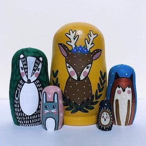 May include: A set of five hand-painted wooden nesting dolls. The largest doll is yellow with a deer design, the next is green with a badger design, the next is pink with a rabbit design, the next is brown with a fox design, and the smallest is gold with a bird design.