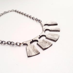 May include: A silver-toned necklace with a chain and a pendant featuring five textured, rectangular shapes. The necklace is displayed on a white surface, highlighting the contrast of the metal.