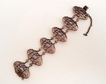 Pode incluir: Uma pulseira de cobre com fecho de corrente. A pulseira é feita de uma série de elos em forma de coração conectados, cada um com um delicado design de filigrana.