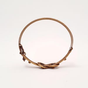 Może przedstawiać: Okrągła bransoletka typu bangle z metalu w kolorze brązu, z teksturowanym, skręconym węzłem u podstawy. Bransoletka posiada mały element w postaci łańcuszka oraz zawiasowy mechanizm zapięcia z jednej strony, wykończony w metalicznym odcieniu brązu.