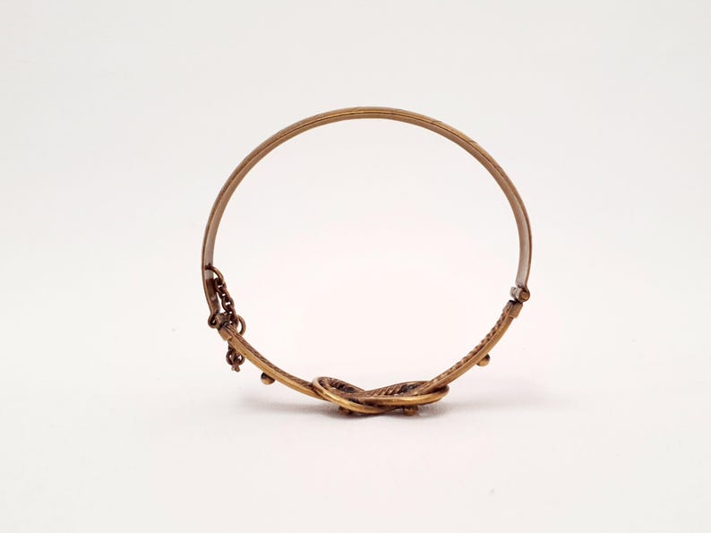 Może przedstawiać: Okrągła bransoletka typu bangle z metalu w kolorze brązu, z teksturowanym, skręconym węzłem u podstawy. Bransoletka posiada mały element w postaci łańcuszka oraz zawiasowy mechanizm zapięcia z jednej strony, wykończony w metalicznym odcieniu brązu.