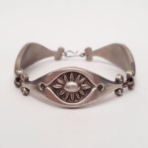 May include: Silver-toned bracelet featuring a central oval design with a detailed sunburst pattern. The bracelet has curved, elongated links and a simple clasp. The metal has a slightly aged appearance, suggesting a vintage or antique style.