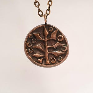 Peut inclure: Un collier avec pendentif de couleur bronze. Le pendentif rond présente un motif d'arbre stylisé en relief avec des accents circulaires. La chaîne du collier est également de couleur bronze et est suspendue sur un fond blanc.