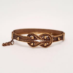 Może przedstawiać: Bransoletka typu bangle z zawiasem, wykonana z metalu w kolorze brązu, z centralnym motywem węzła nieskończoności. Obręcz składa się ze skręconych drucik&oacute;w ozdobionych małymi, gładkimi, kulistymi nitami. Z jednej strony przymocowany jest kr&oacute;tki łańcuszek zabezpieczający.