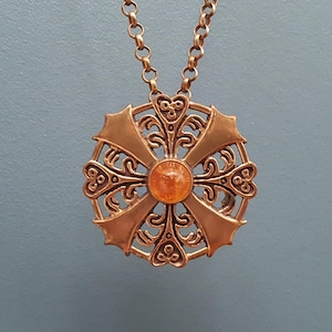 Puede incluir: Un collar dorado con un gran colgante adornado con un diseño de cruz y una piedra preciosa naranja en el centro.