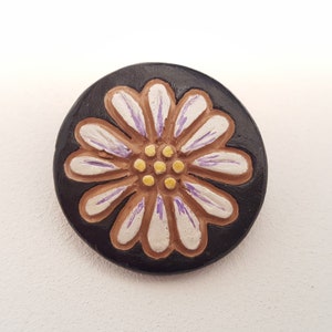 May include: A round, black ceramic button featuring a white daisy flower design. The flower has a brown centre with yellow dots and purple accents on the petals.