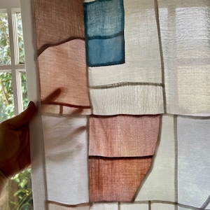 Pode incluir: Uma cortina de tecido branco com um design de patchwork. O tecido é tingido em tons de marrom, azul e branco. A cortina está pendurada em uma janela.
