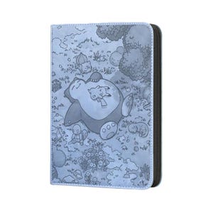 May include: A light blue card binder featuring a detailed illustration of a sleeping Snorlax with Pikachu and other Pok&eacute;mon characters in a grassy landscape. The binder has a black spine and a zipper closure.