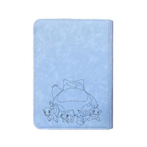 May include: A light blue card binder featuring a line drawing of Snorlax and other Pok&eacute;mon characters. The binder has a textured surface and a stitched border. The design includes Bulbasaur, Pikachu, and Squirtle.