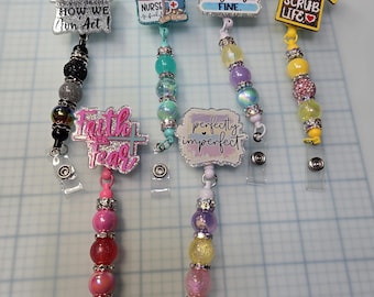 May include: A collection of badge reels with various designs and phrases. The reels feature phrases like "I Match Energy," "Nurse," "It's Fine," and "Scrub Life." Each reel has a unique beaded design in colors like pink, purple, and yellow. The reels are designed for healthcare professionals.