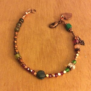Puede incluir: Un brazalete de cobre con cuentas verdes, rosas y marrones, un dije en forma de corazón y un pequeño dije de cobre con un diseño de hoja.
