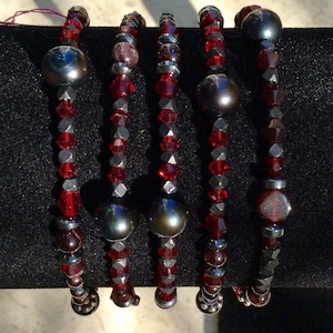 Op de afbeelding: Vijf armbanden met rode, zilveren en donkergrijze kralen. Elke armband heeft een centrale grote, donkergrijze kraal, omringd door kleinere rode en zilveren kralen. De armbanden worden gepresenteerd op een zwart oppervlak.