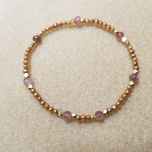 Op de afbeelding: Een delicate armband met kleine, perzikroze parels afgewisseld met facetgeslepen goudkleurige kralen en ronde, lichtpaarse edelstenen. De armband is cirkelvormig.