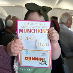 May include: A child holds a sign that reads "THANK YOU FOR BEING PART OF OUR MUNCHKIN'S first flight" with a Dunkin' logo. The sign has a pink border and a snowflake and Christmas tree graphic.