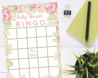 May include: A baby shower bingo card with a floral design in pink and green. The card has a grid of squares with the words "Baby Shower BINGO" at the top and "Free Space" in the center. A black pen, a green notebook, a binder clip, and a small potted plant are also visible.