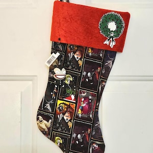 May include: A Christmas stocking featuring a red cuff with a green wreath and skull design. The body of the stocking is black with various character illustrations from The Nightmare Before Christmas.
