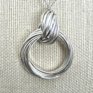 May include: Silver-toned necklace featuring a large, textured pendant with a knotted design. The pendant is composed of multiple intertwined rings, creating a layered effect. The chain is delicate and complements the bold pendant.