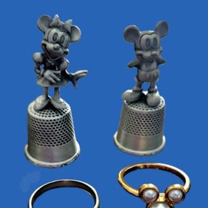 May include: Two silver-coloured figurines of cartoon mice on thimbles, and two rings. One ring is gold-coloured with three pearls, and the other is silver-coloured with a Mickey Mouse design. The background is blue.