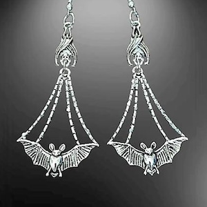 May include: A pair of silver dangle earrings featuring a bat design. The earrings have a dangling bat charm with a delicate chain and a bat-shaped pendant.