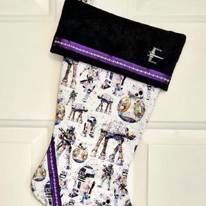 May include: A Star Wars-themed Christmas stocking with a black velvet cuff and purple ribbon trim. The body of the stocking features a white background with colorful illustrations of various Star Wars characters and vehicles.