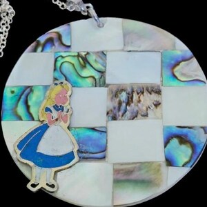 May include: A silver-toned pendant necklace featuring a round, checkered design with mother-of-pearl and abalone shell squares. A cartoon figure of Alice in Wonderland is on the pendant. The necklace has a silver chain.