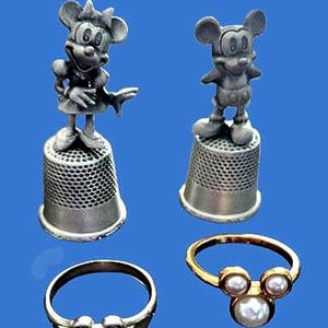 Peut inclure: Deux dés à coudre en métal argenté surmontés de figurines sculptées représentant un personnage de souris, accompagnés de deux bagues : une bague en métal argenté et une bague en métal doré ornée de trois perles en forme d'oreilles de souris.