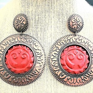 Könnte beinhalten: Ein Paar großer, kreisförmiger Ohrringe mit einem Sonnendesign. Die Ohrringe haben eine rote Sonne in der Mitte, umgeben von einem silberfarbenen Rand mit einem komplizierten Muster. Der obere Teil ist eine kleinere, passende silberne Scheibe.
