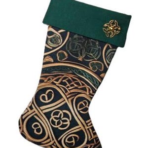 Puede incluir: Un calcetín navideño con cuerpo de tela oscura y un intrincado patrón de nudos entrelazados en dorado y verde. El puño superior es de tela verde oscuro lisa y está adornado con un emblema metálico circular de color dorado en el lado derecho.
