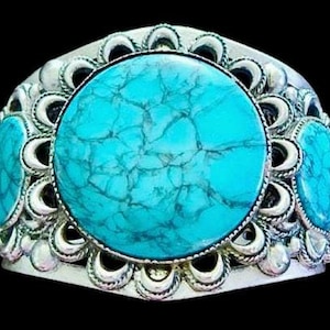 Puede incluir: Un brazalete plateado con tres grandes piedras turquesas ovaladas. El brazalete tiene un diseño ornamentado con un borde festoneado y detalles decorativos alrededor de las piedras. Las piedras turquesas tienen un aspecto moteado.