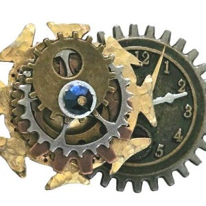 May include: Steampunk-style brooch featuring a clock face and gears in shades of gold, silver, and bronze. The brooch has a central blue gem and is designed with intricate mechanical details. The clock face shows the numbers 1 through 5.