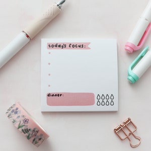 May include: A pink and white notepad with the text "today's focus:" and "dinner:" printed on it. The notepad has a pink and white striped design and a row of raindrops at the bottom.