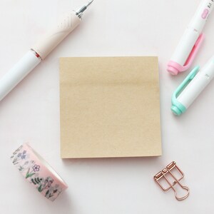 May include: A brown paper note pad with a pink floral washi tape, a pink highlighter, a light blue highlighter, and a gold binder clip on a white surface.