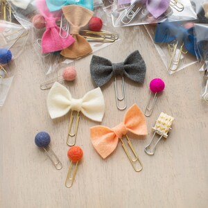 May include: Assortment of colourful felt bow paper clips with gold metal clips. The bows are in various colours, including pink, white, grey, and peach. There are also small felt balls in various colours, including pink, blue, and orange.