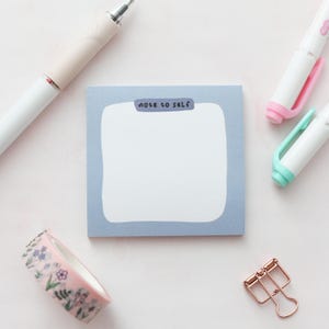 May include: A blue notepad with the words "note to self" printed on the top. The notepad is surrounded by various stationery items, including a pink and white pen, a blue and white pen, a roll of washi tape with floral designs, and a gold paper clip.