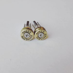 Może przedstawiać: Para złotych spinek do mankietów w kształcie łusek po nabojach 9 mm Luger. Łuski są grawerowane napisem "SPEEER 9mm LUGER".