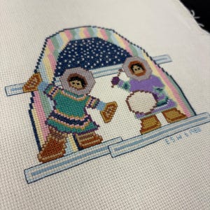 May include: Cross-stitch embroidery featuring two figures in winter clothing under a rainbow-colored arch. The figures are wearing parkas and boots. The background is a starry night. The text "ESW 6 1986" is visible.
