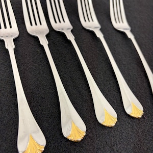 May include: A set of six silver forks with a gold-colored floral design at the base of the handle. The forks are arranged diagonally against a dark background, showcasing their elegant design.