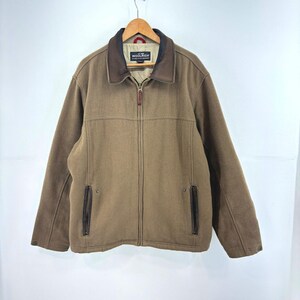 May include: A tan zip-up jacket featuring a dark brown collar, long sleeves, and two vertical zippered pockets on the front. The interior label displays the text Woolrich. The jacket is displayed on a wooden hanger against a plain background.