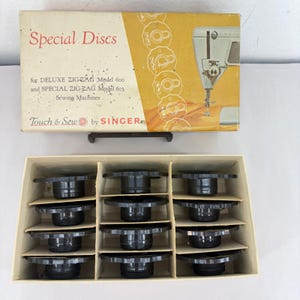 May include: A vintage Singer Touch & Sew box containing twelve black plastic special discs for Deluxe Zig-Zag Model 600 and Special Zig-Zag Model 603 sewing machines. The box lid features red and black text with an illustration of a sewing machine.