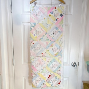May include: A rectangular patchwork quilt featuring a repeating pattern of pastel-colored squares in shades of pink, yellow, blue, and white. The fabric squares are decorated with small illustrations of cartoon animals and floral motifs.