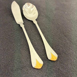 May include: A butter knife and spoon set. The silver-colored utensils have a gold-colored, fan-shaped design at the base of the handles. The spoon has a rounded bowl. The knife has a flat, rounded blade.
