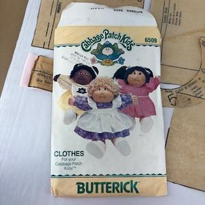 May include: A Butterick 6509 sewing pattern envelope for Cabbage Patch Kids clothes. The front features three dolls wearing different outfits, including a purple dress with a white apron, a yellow dress, and a pink patterned dress. Text includes Cabbage Patch Kids, 6509, and Butterick.