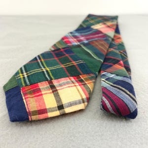 May include: A necktie constructed from multiple patchwork sections of cotton fabric featuring various plaid and tartan patterns in shades of green, red, yellow, blue, and black. The tie displays a mix of diagonal and straight fabric orientations.